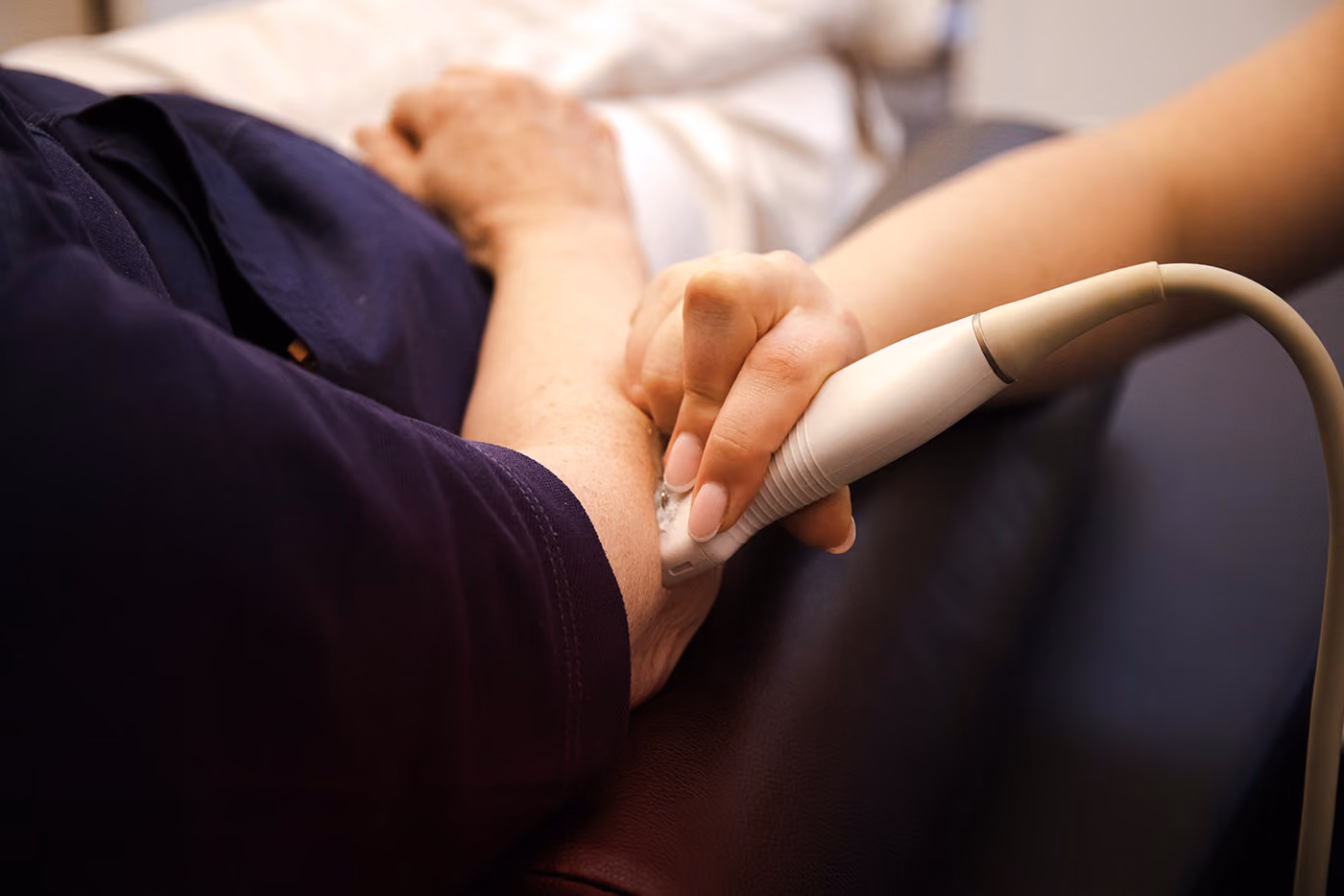 Een hand houdt een witte ultrasoundtransducer bij de pols van een persoon die een donkerblauwe mouw draagt, tijdens een medische onderzoekssituatie.