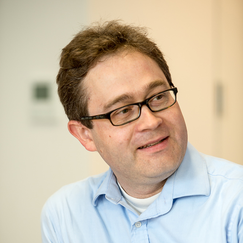Man with short curly brown hair and glasses, wearing a light blue collared shirt, looking slightly to his right with a faint smile.