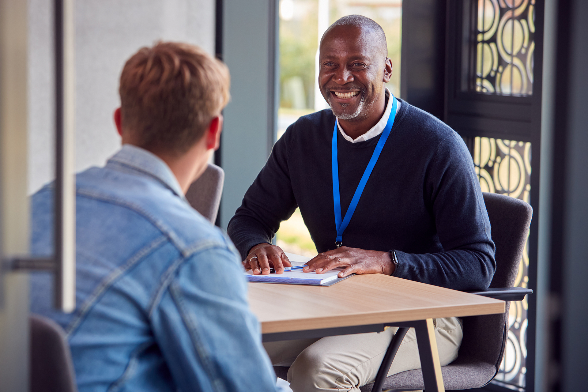 Smiling man in a black sweater with a blue lanyard sitting at a table, talking to a person in a denim jacket across from him.