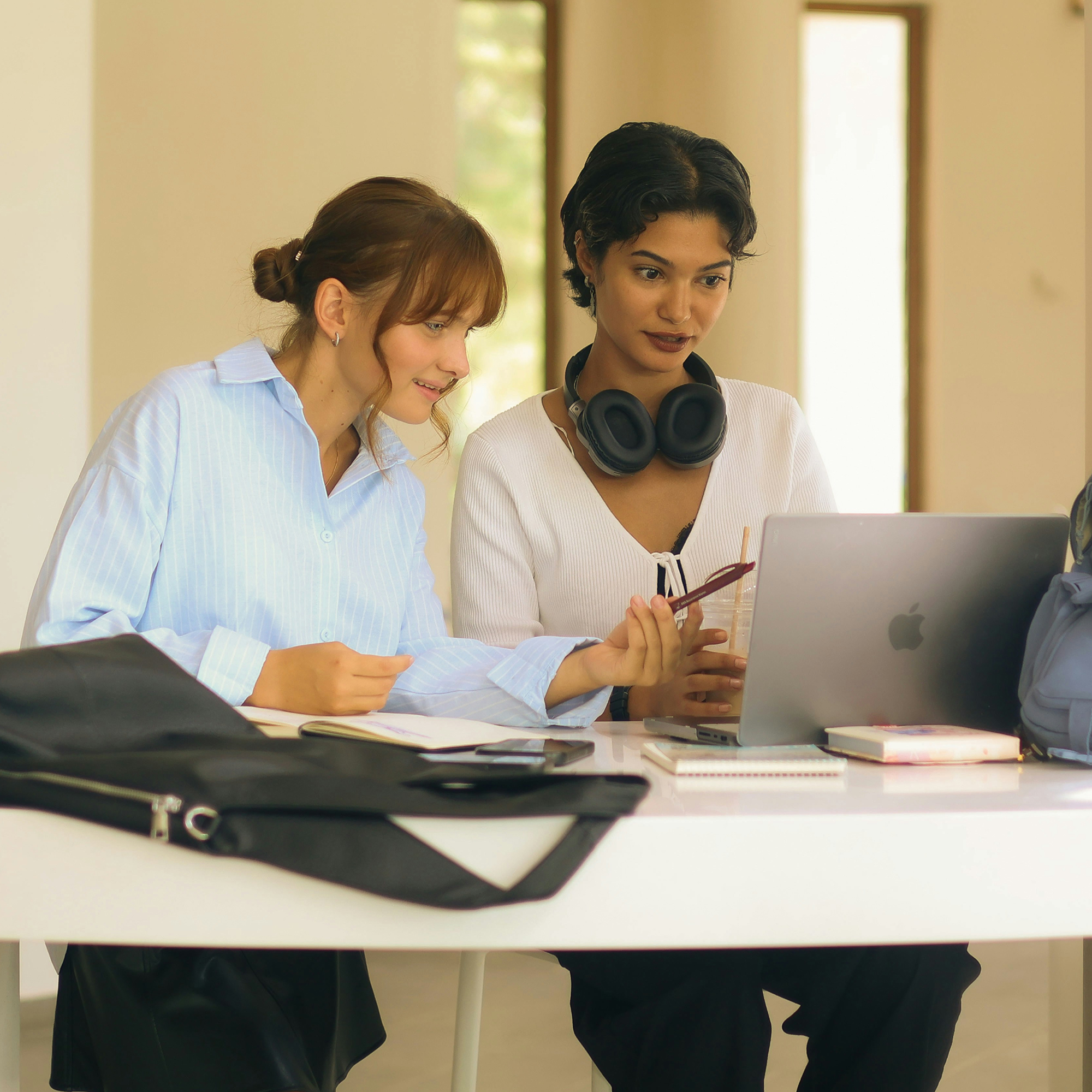 Two young women sitting at a white table working together on a laptop in a bright room, one holding a pen and notebook while the other wears headphones around her neck and holds a drink.