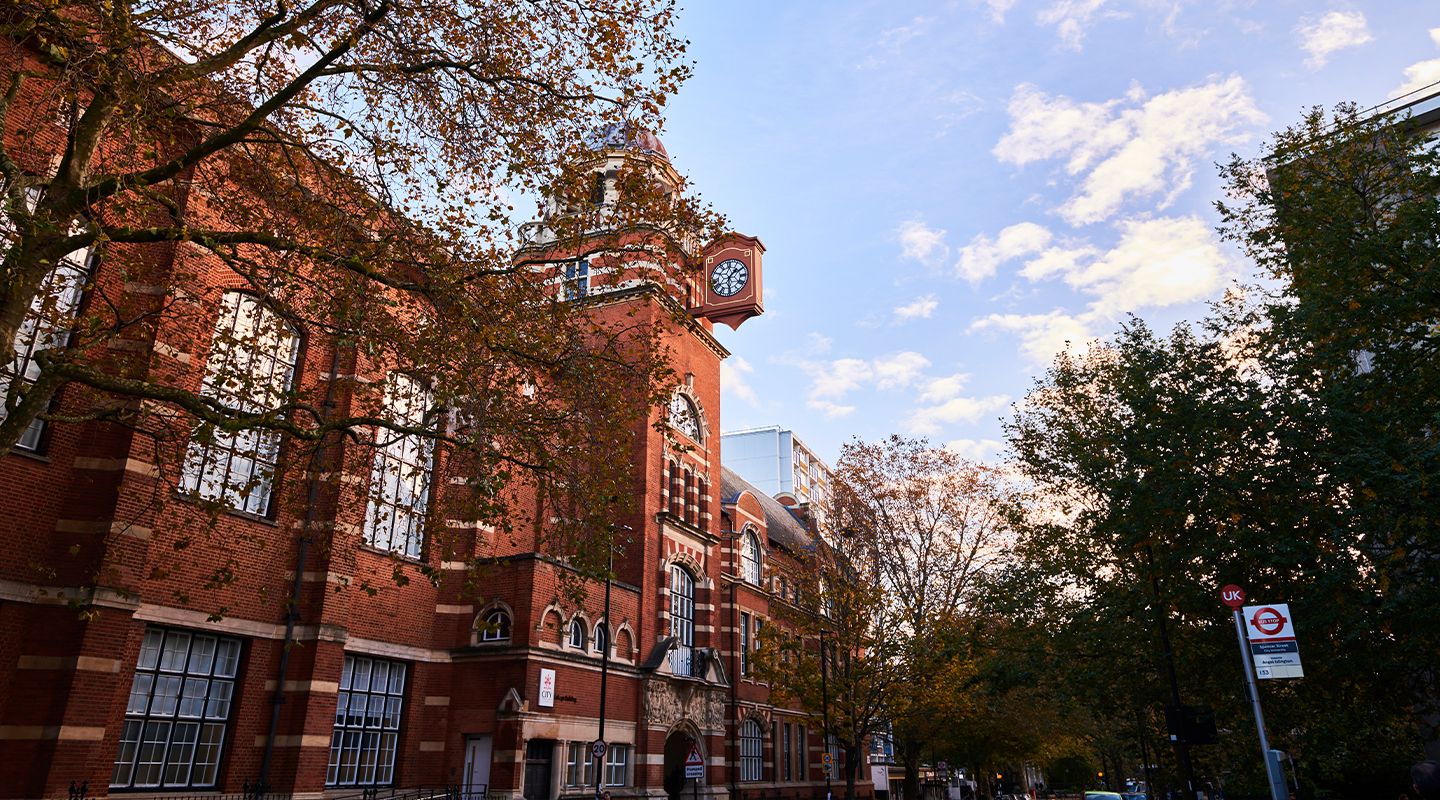 Red-brick building with large windows and a clock tower, surrounded by autumn trees under a partly cloudy blue sky.