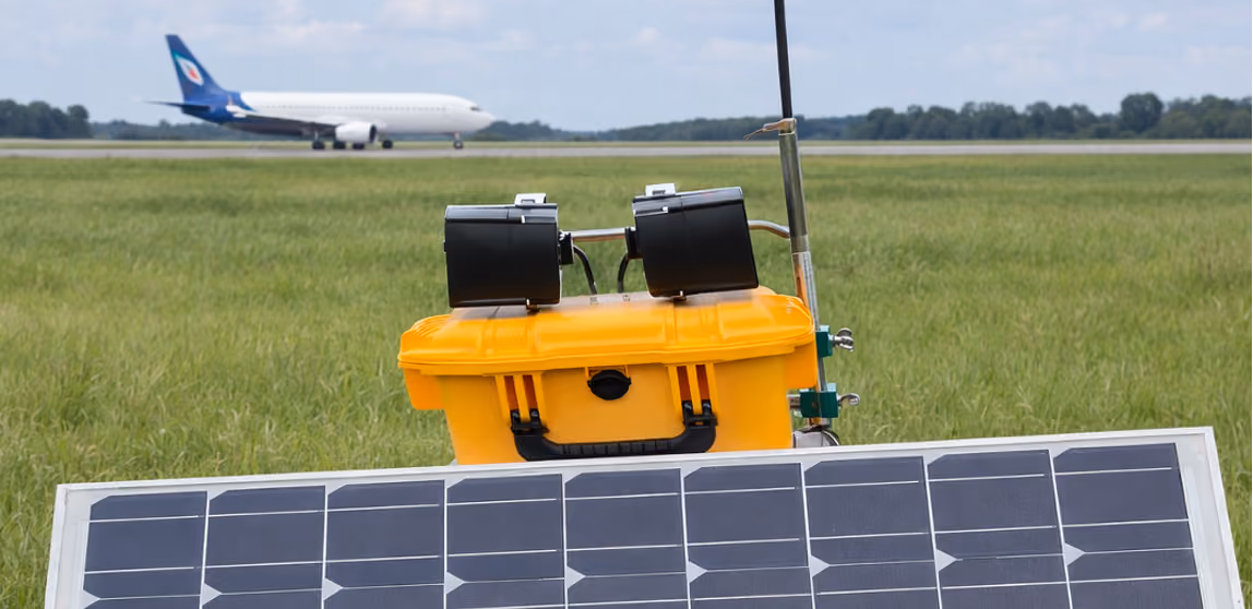 Yellow weather monitoring device with solar panels on grass near airport runway with plane in background.