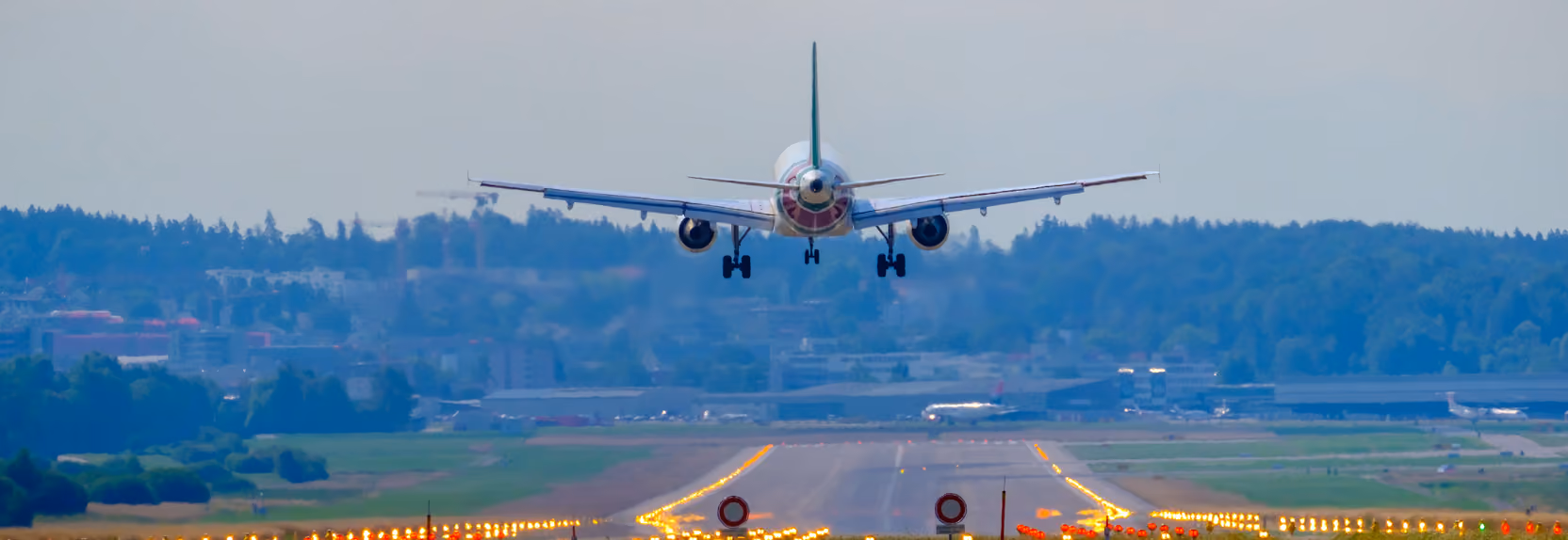 Commercial airplane descending to land on runway with forest and buildings in the background.
