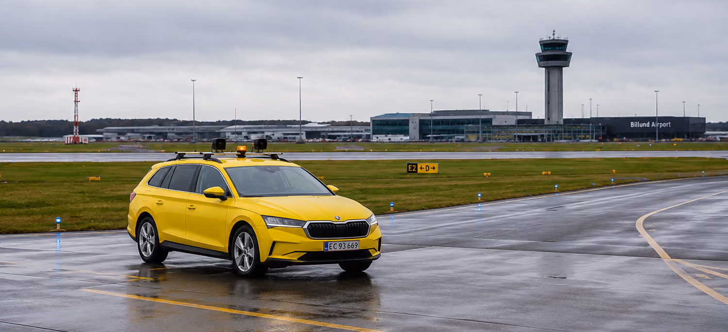 Yellow car with rooftop sensors driving on wet runway at Billund Airport with control tower in background.