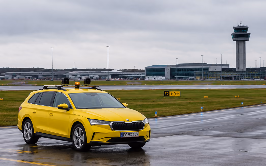 Yellow airport service car on wet runway with control tower and terminal in background on cloudy day.