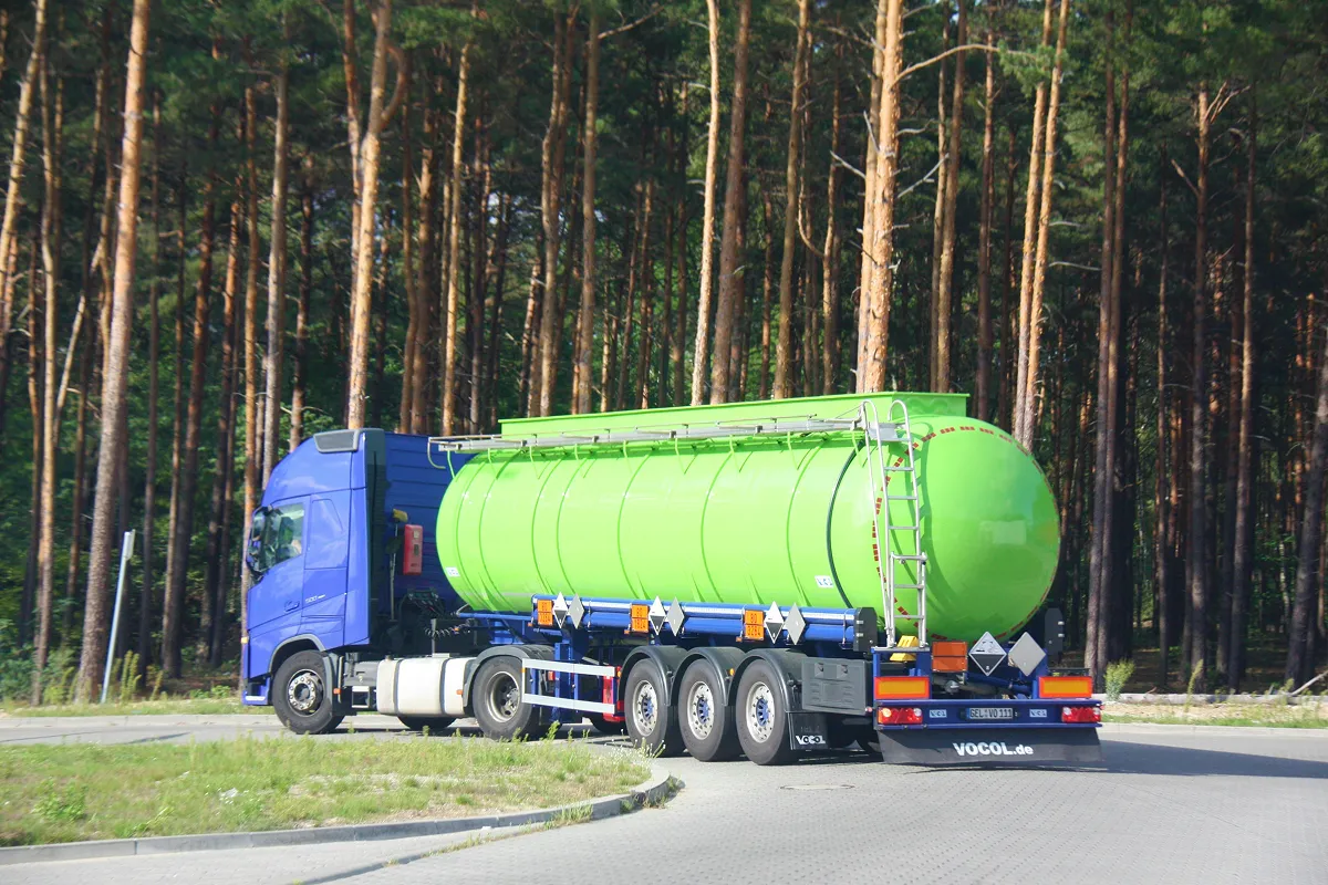 Blue truck with a large bright green fuel tanker trailer on a paved road near a pine forest.