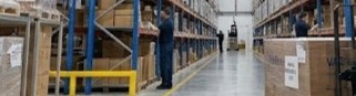 Warehouse aisle with tall shelves stocked with boxes and a worker walking along the aisle.