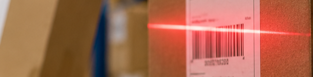 Red laser scanning a barcode on a cardboard box in a warehouse or shipping area.