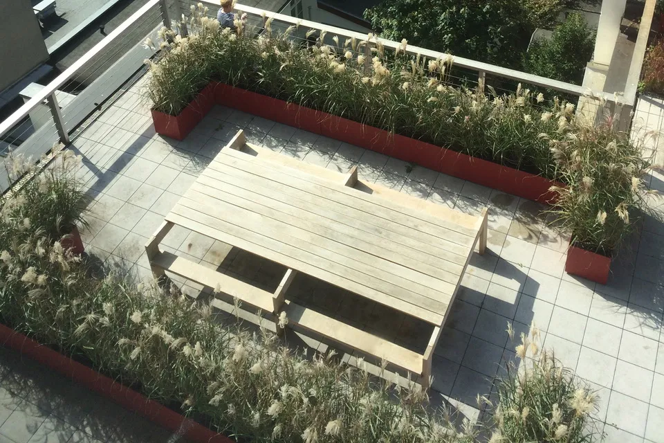Roof terrace with a large light wooden table and two benches, surrounded by rectangular red planters with tall, delicate grasses, on gray square floor tiles.