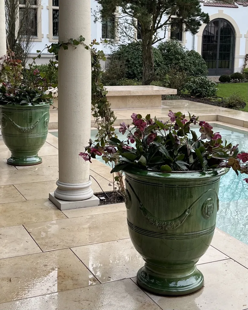 Green decorative flower pot with blooming pink and purple flowers on a stone floor next to a white column and a small water basin in a garden with shrubs and a white building in the background.