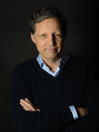 Portrait of a middle-aged man wearing a dark blue sweater over a light blue shirt against a black background, arms crossed, slight smile.