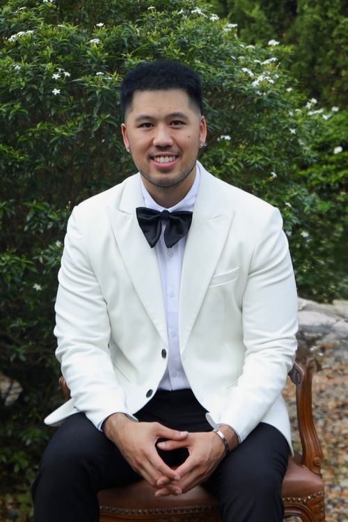 Smiling man in white tuxedo jacket with black bow tie sitting on a chair outdoors
