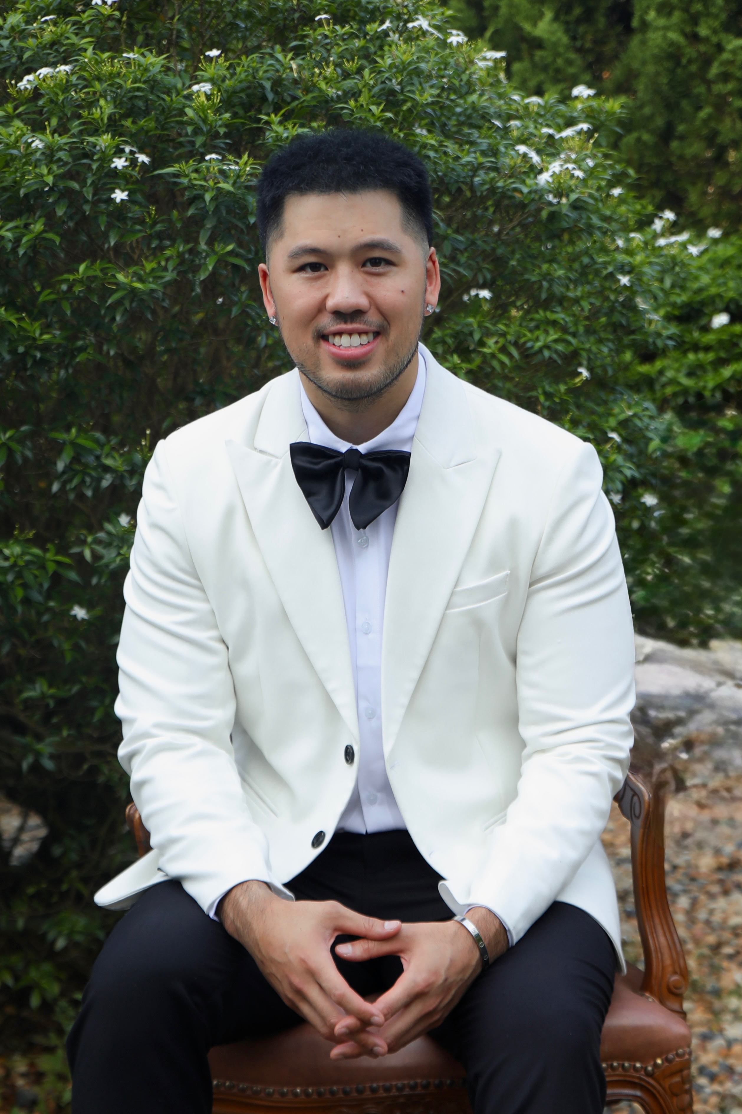 Smiling man in white tuxedo jacket with black bow tie sitting on a chair outdoors