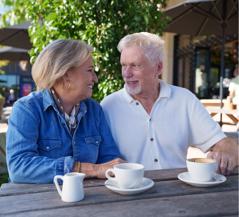 Smiling elderly couple sitting at wooden table outdoors with coffee cups, enjoying conversation.