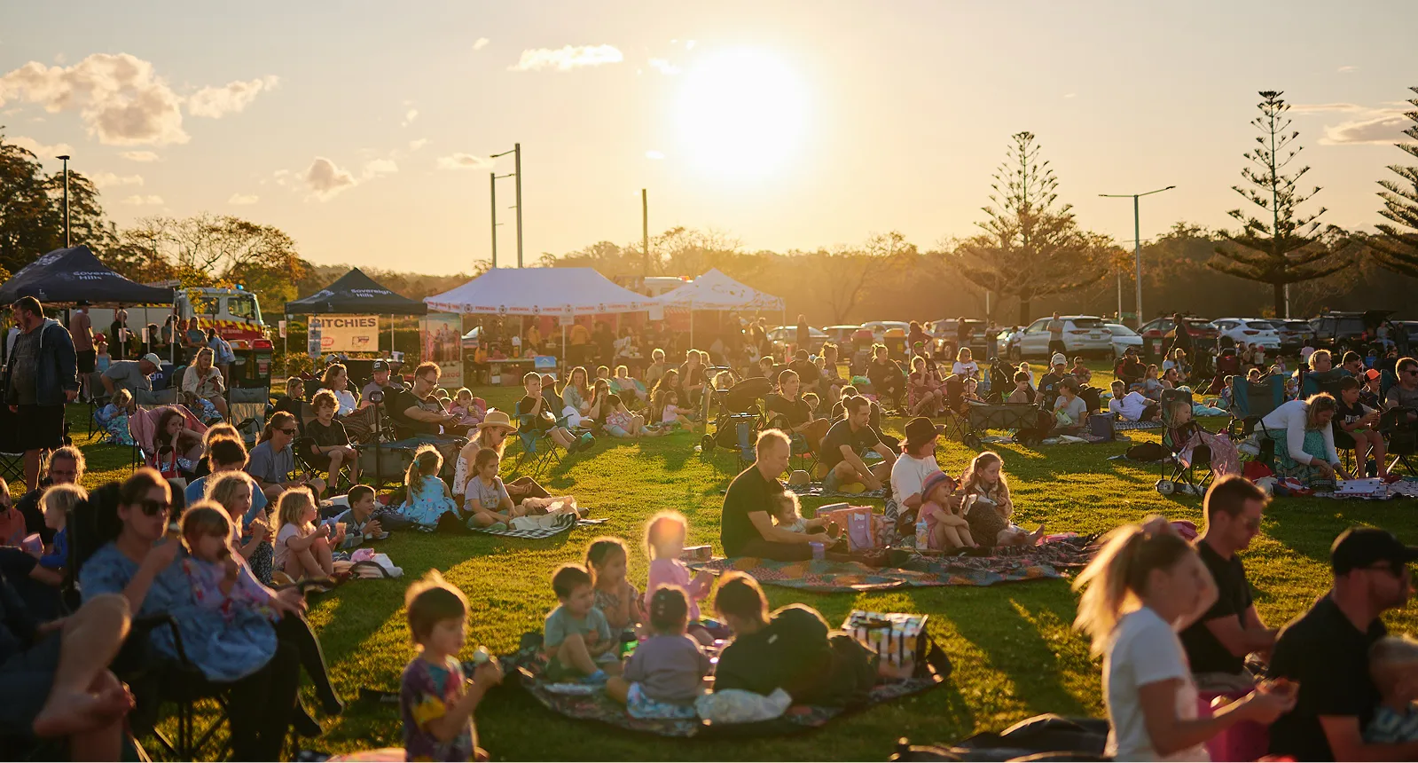 People gathered on grass at an outdoor event in late afternoon sunlight with tents and trees in the background.