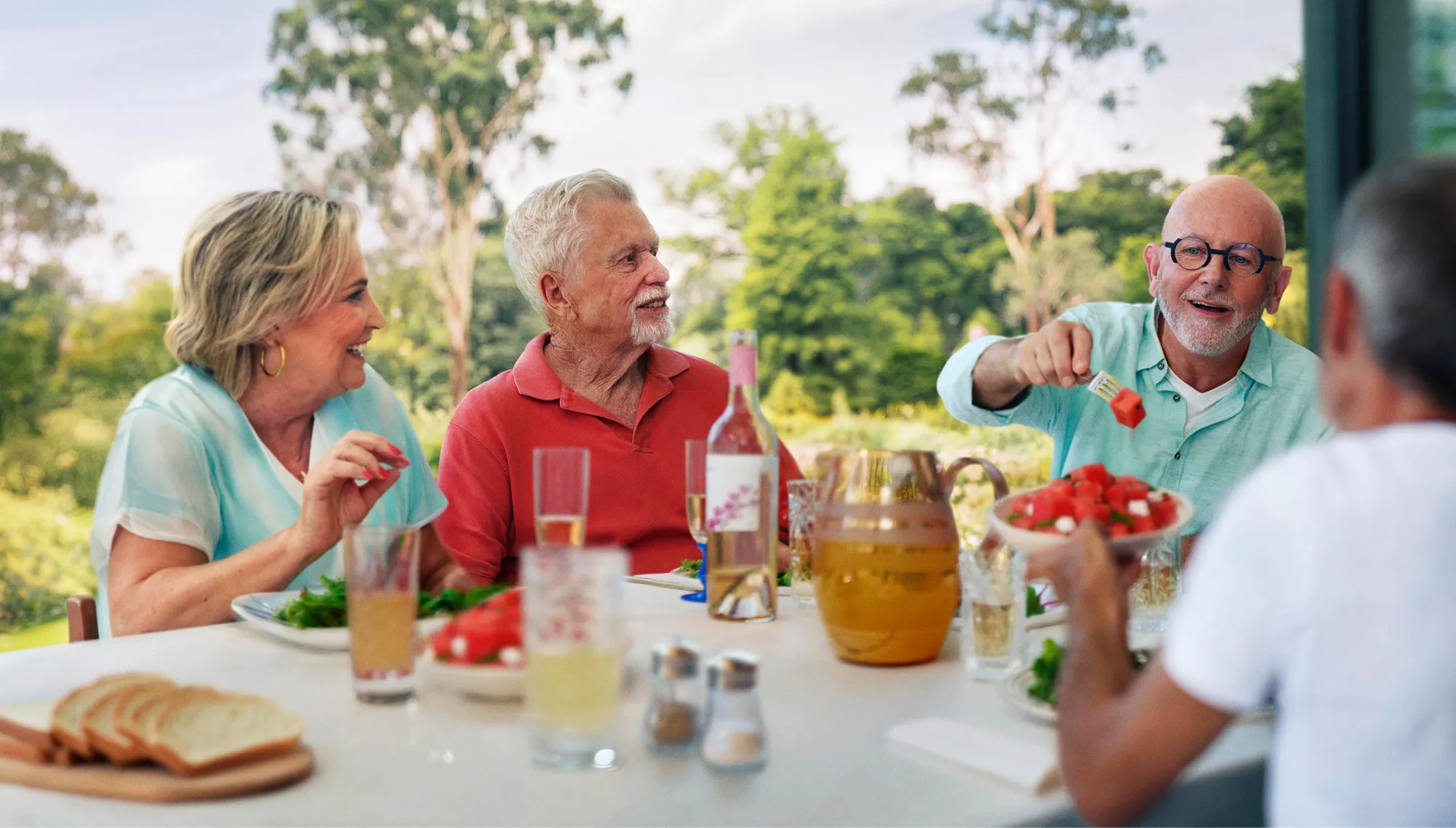 Four older adults enjoying an outdoor meal with drinks and watermelon salad on the table.