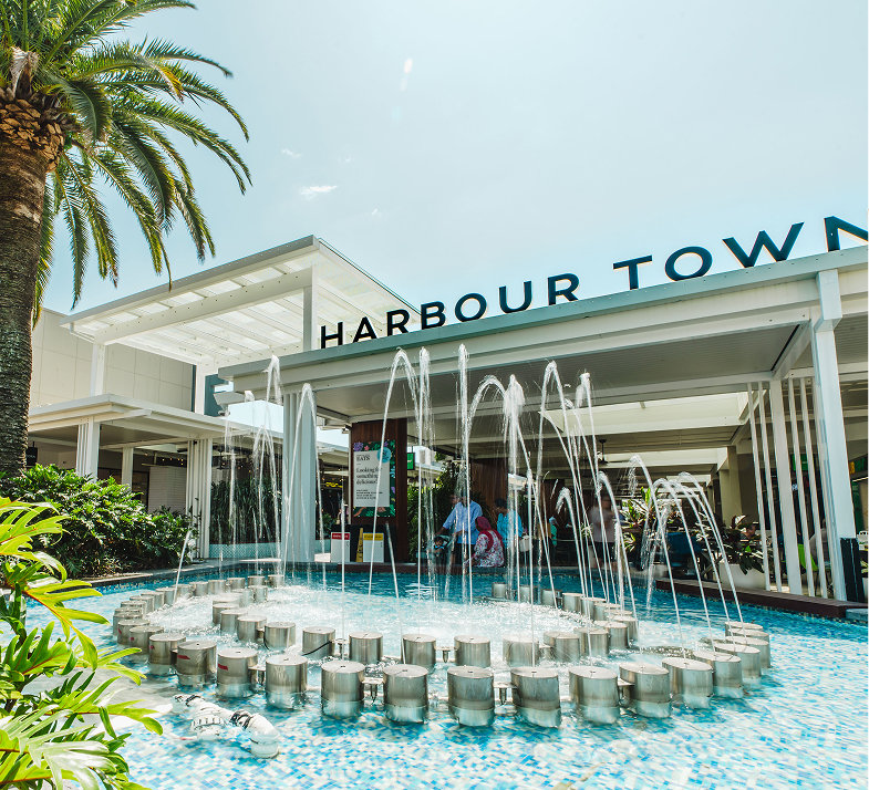 Fountain with water jets in a blue tiled pool in front of white Harbour Town shopping area under clear sky.
