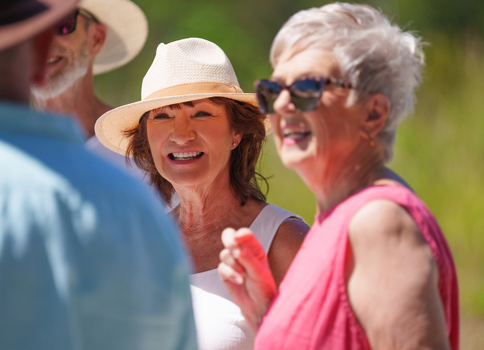 Smiling middle-aged woman in white hat talking with friends outdoors on a sunny day.