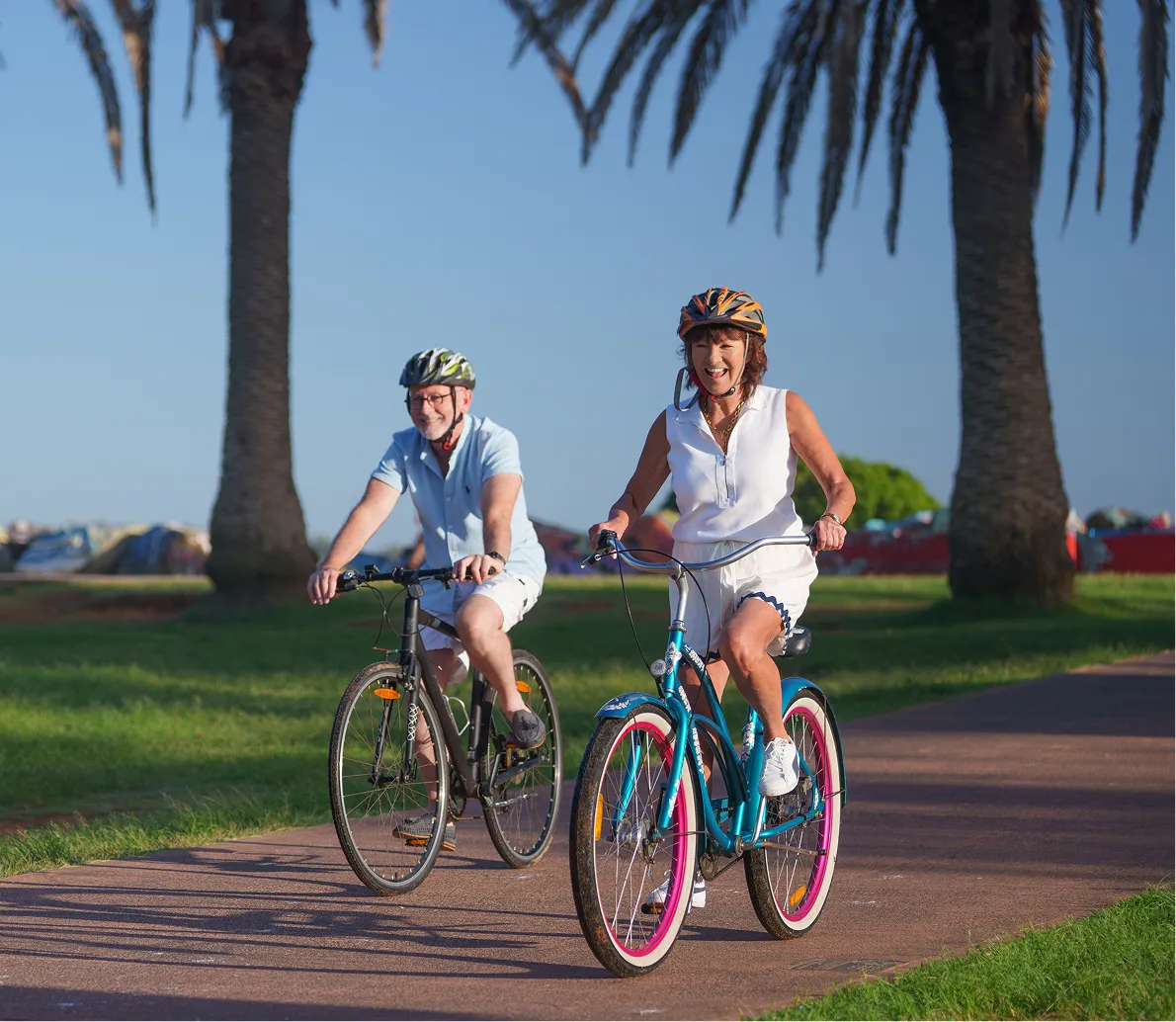 Smiling elderly man and woman wearing helmets riding bikes on a path with palm trees and grass.