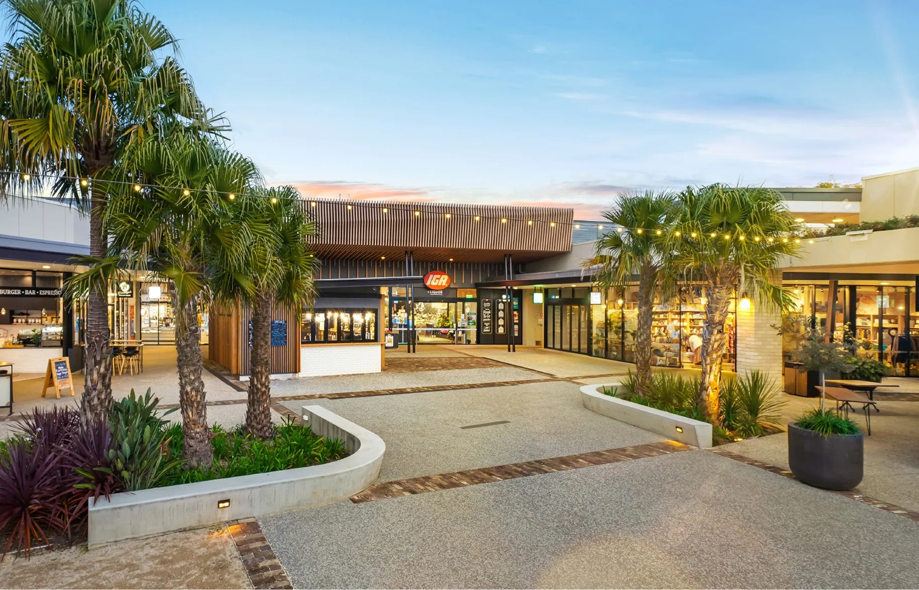 Outdoor shopping area with palm trees, string lights, and an IGA supermarket at sunset.