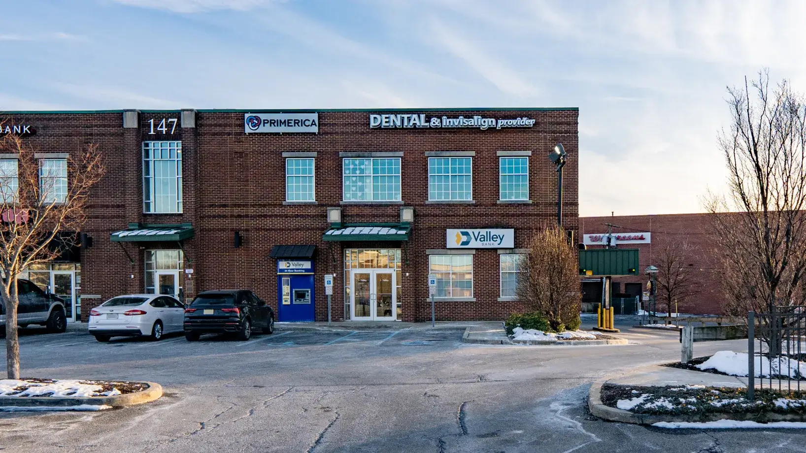 Brick commercial building with signs for Primerica, dental and Invisalign provider, and Valley Bank, with parked cars and patches of snow in the parking lot.