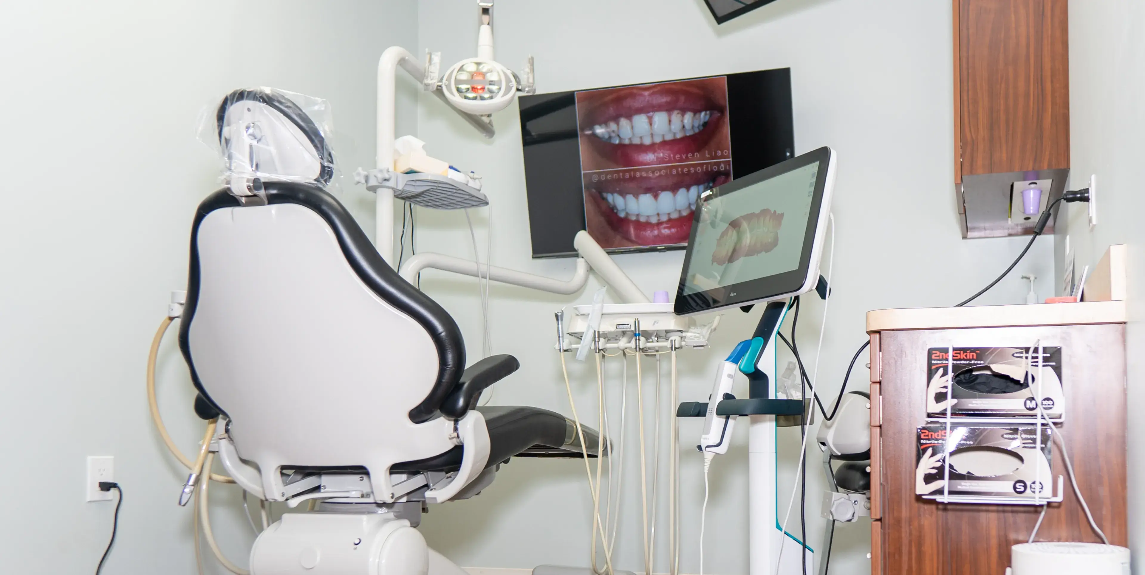 Modern dental chair in a clean dental office with dental tools, a monitor showing a 3D tooth model, and a TV screen displaying before and after teeth whitening images.