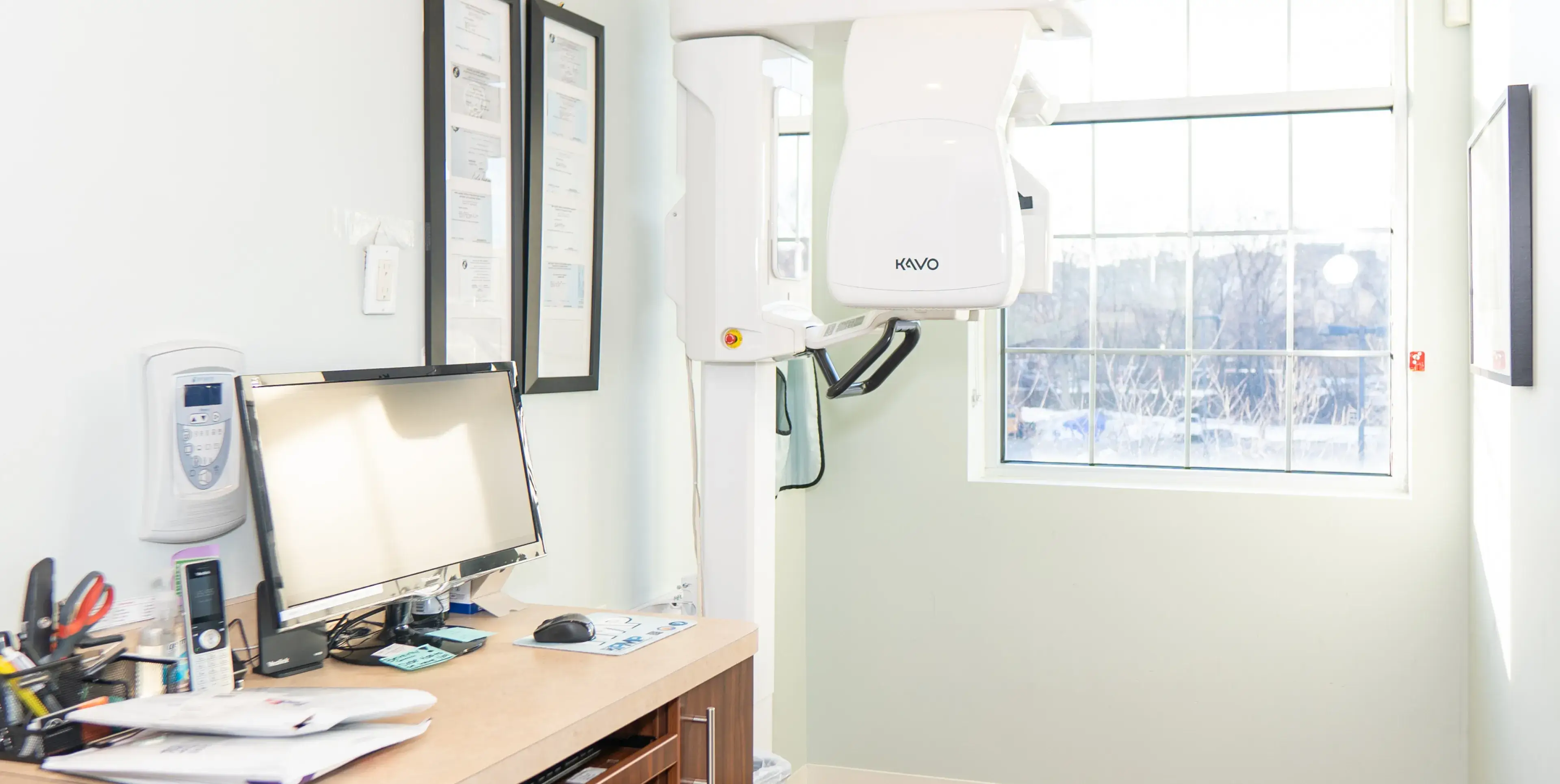 A dental X-ray machine is positioned next to a desk with a computer in a bright room.