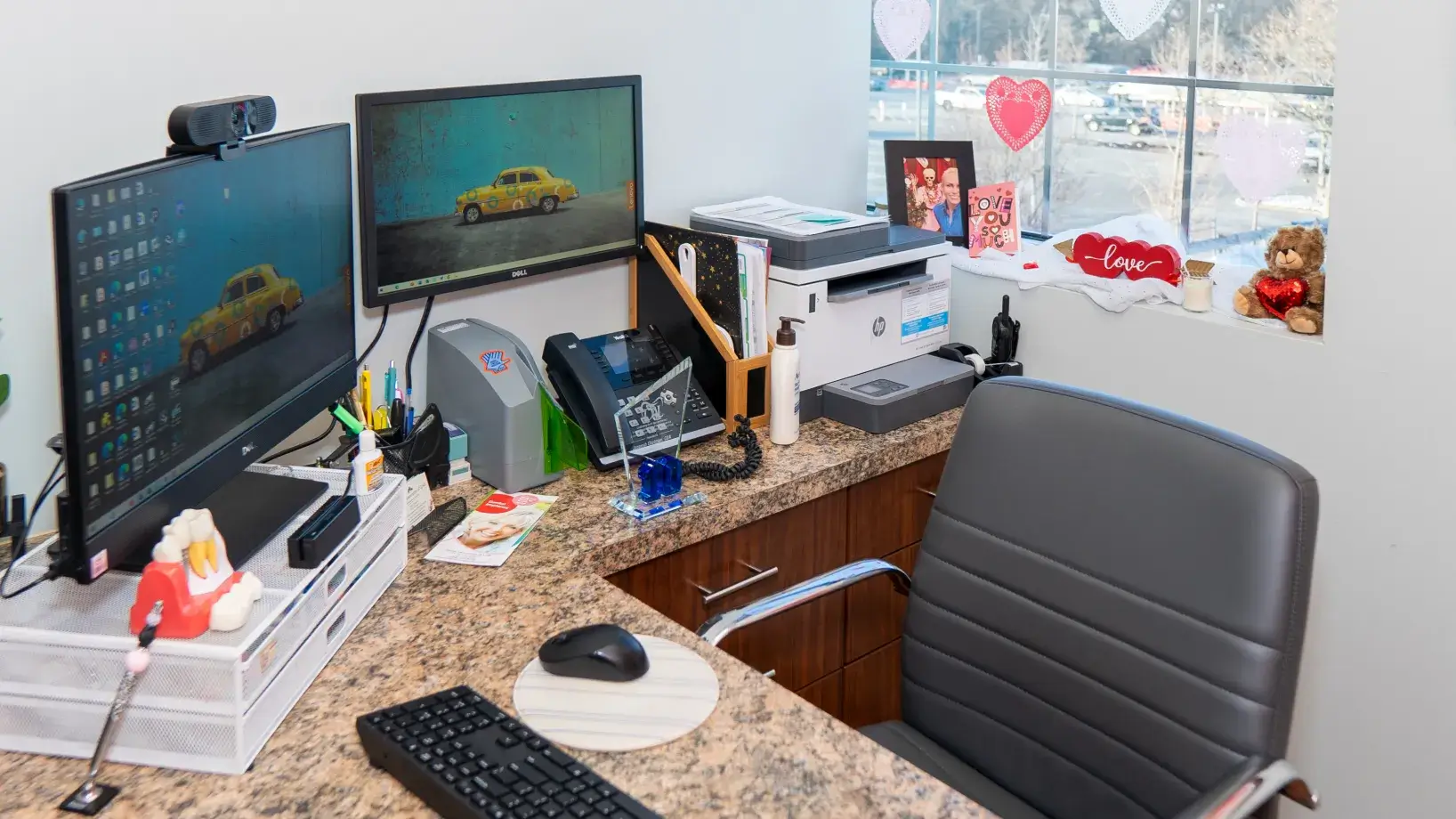 Office desk with two computer monitors, printer, and various decorations on the window ledge.