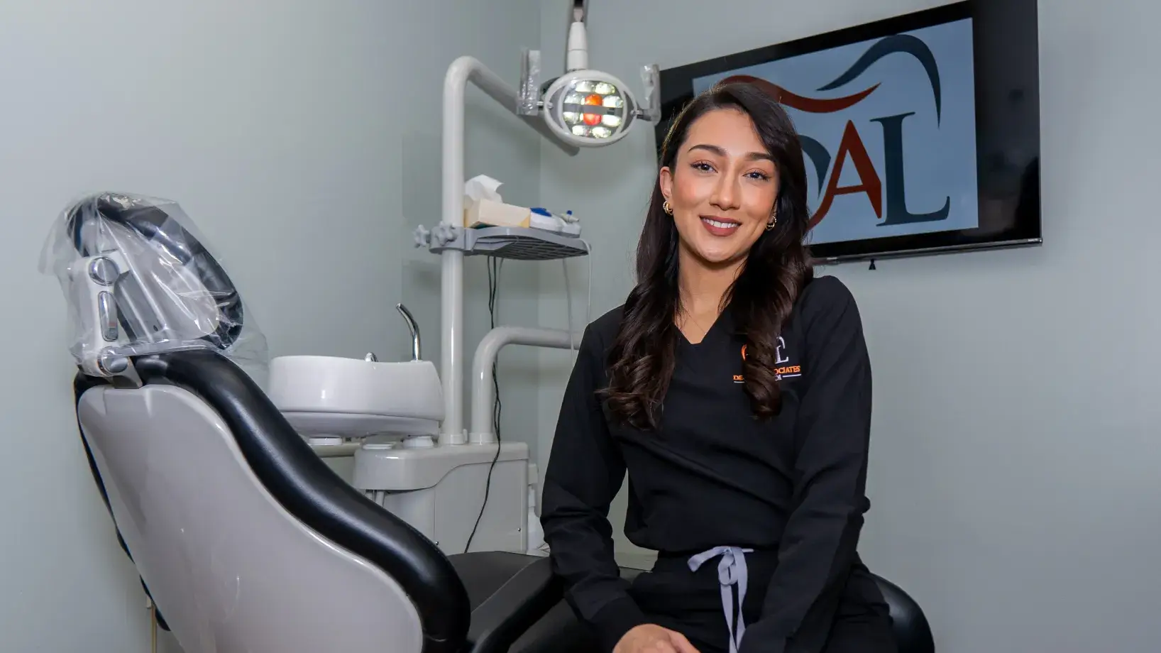 A woman in dental scrubs smiles while sitting in a dental office next to a treatment chair.