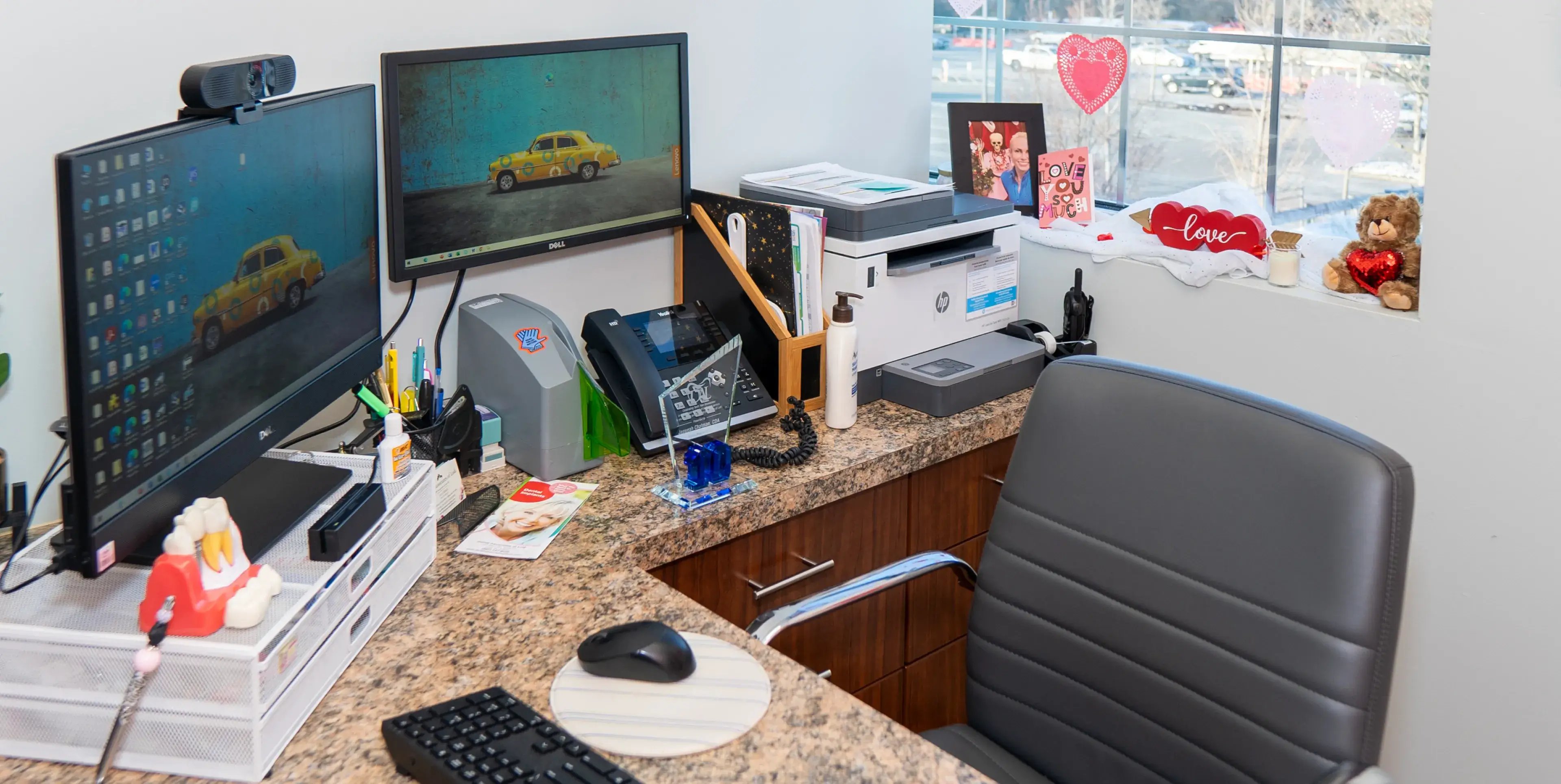 An office desk with dual monitors, a printer, keyboard, mouse, and chair. Decor includes heart-themed items.