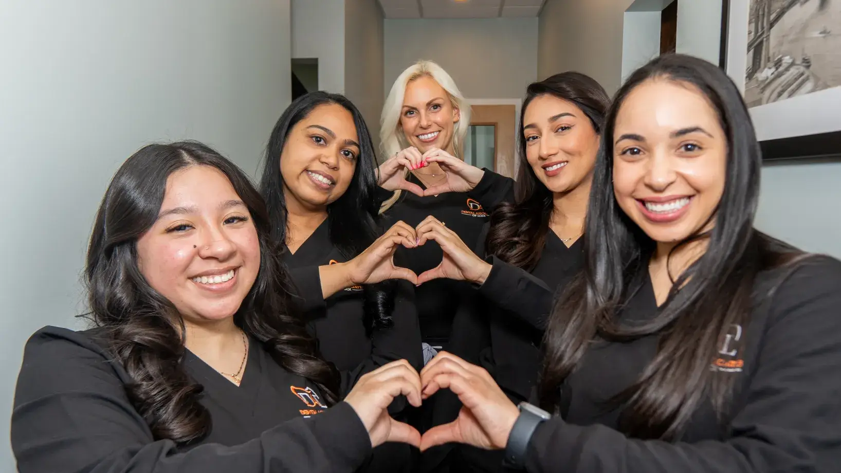 Five women smiling and forming heart shapes with their hands, standing together in a hallway.