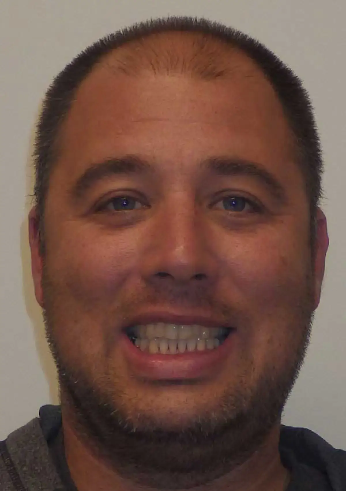 A man with short hair smiling directly at the camera against a plain background.