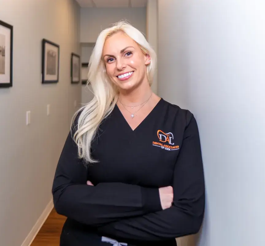 Smiling person with long blonde hair, wearing a black dental uniform, stands in a hallway.