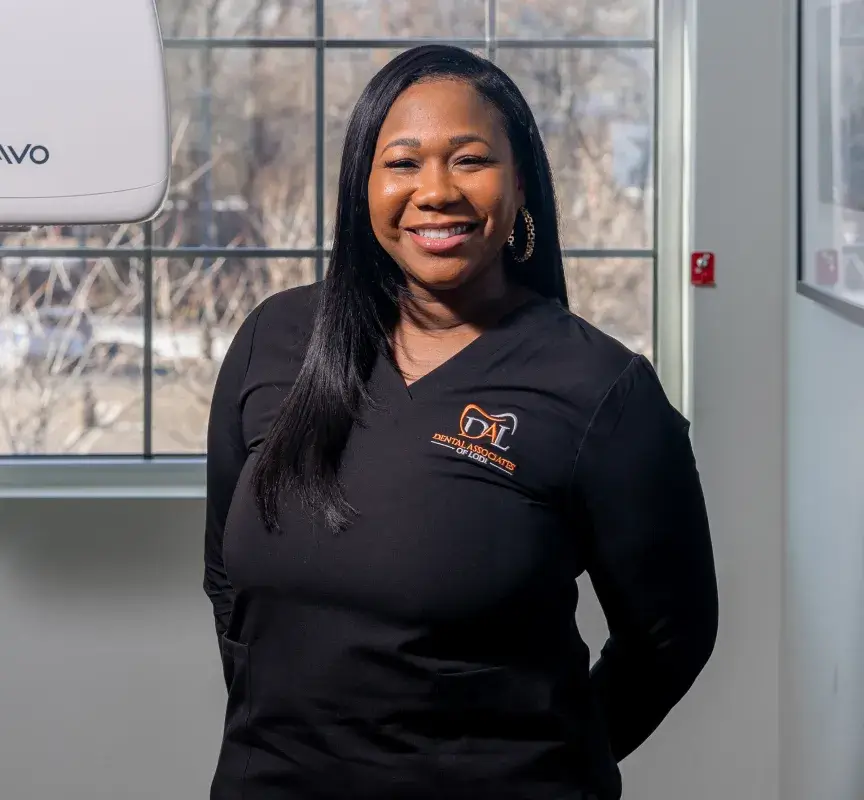 Woman in a dental office, smiling and wearing a black uniform with a dental logo.