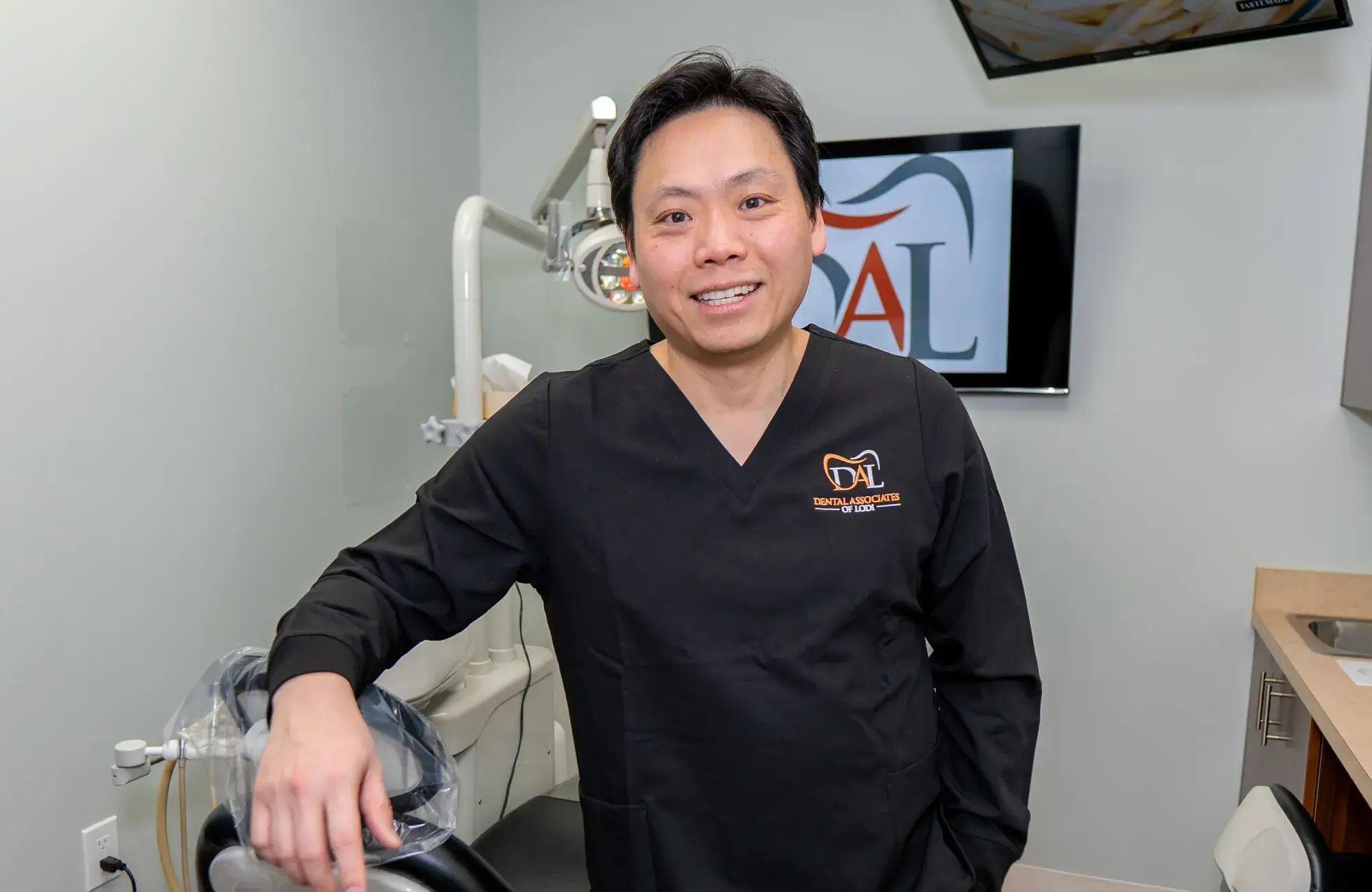 A dentist in a black uniform stands smiling next to a dental chair in the office.