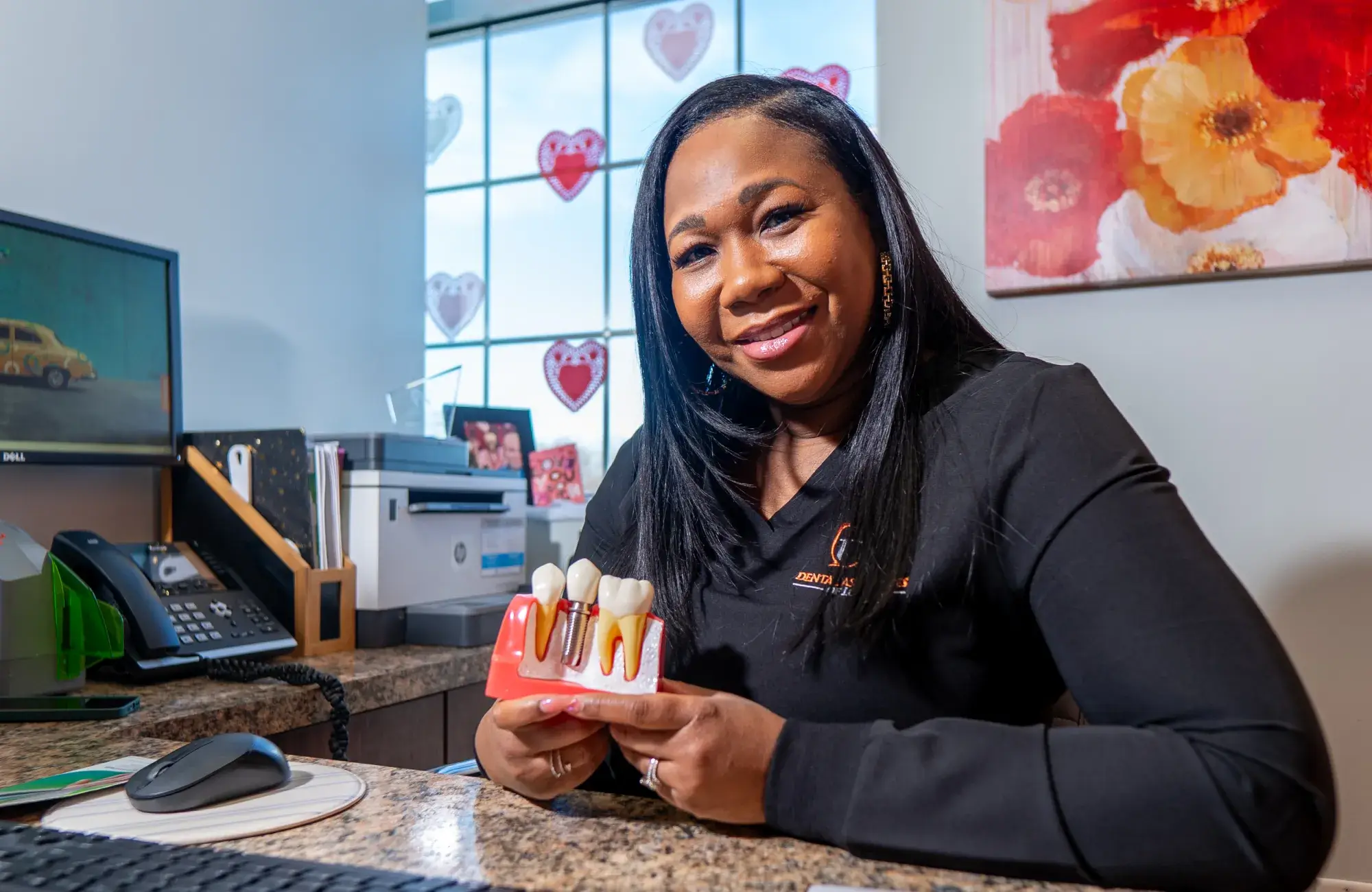 A woman smiles while holding a dental model with teeth sitting at a desk in an office.