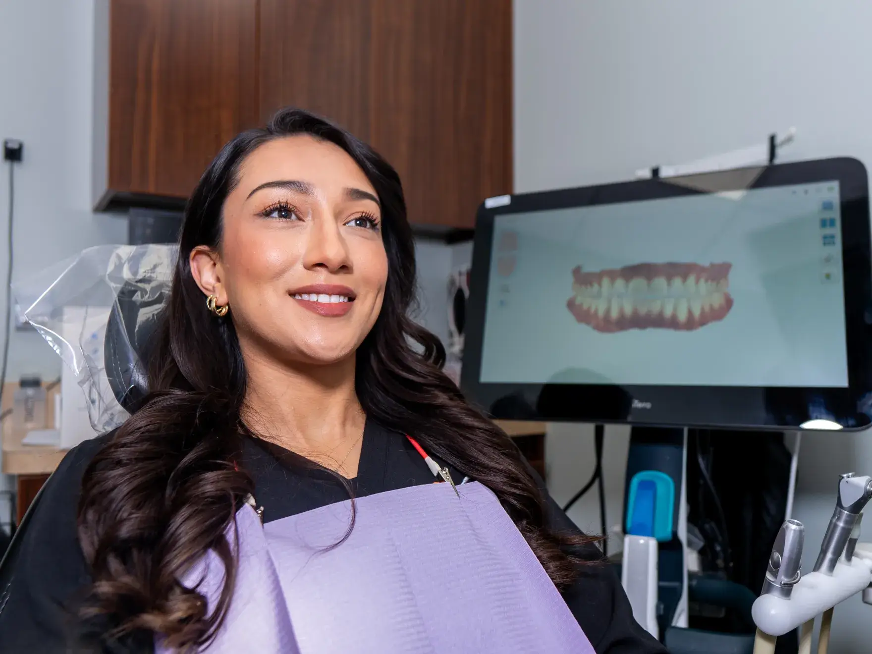 Woman sitting in dental chair, smiling, with dental scan displayed on screen in the background.