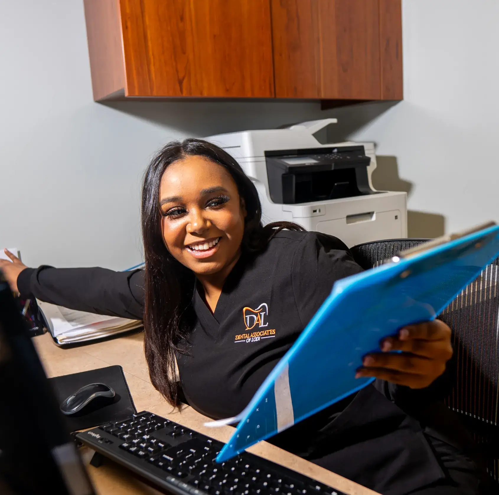 A person in a dental uniform smiles while holding a blue folder at a desk.
