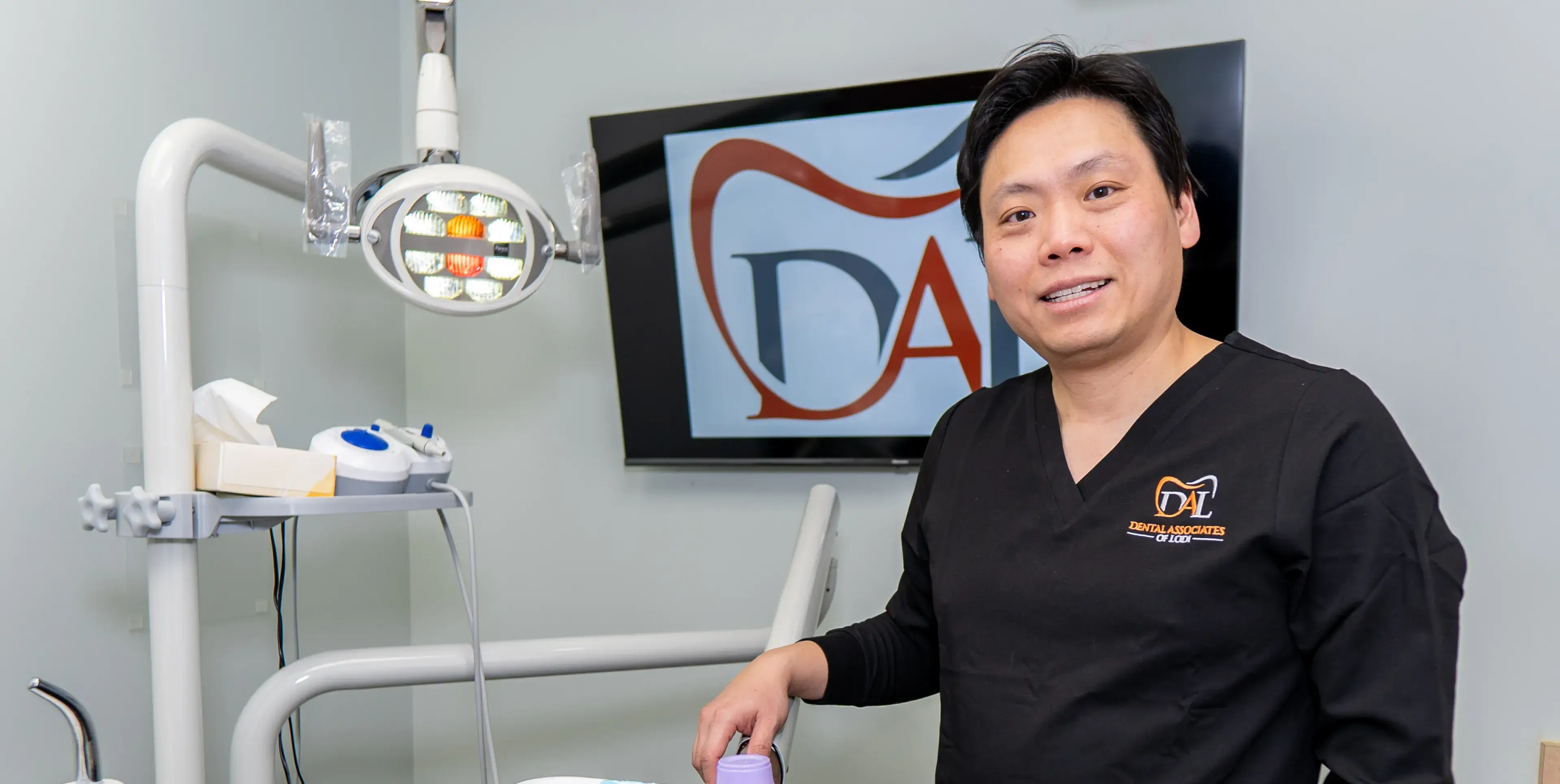 A dentist in scrubs stands beside dental equipment in an office.