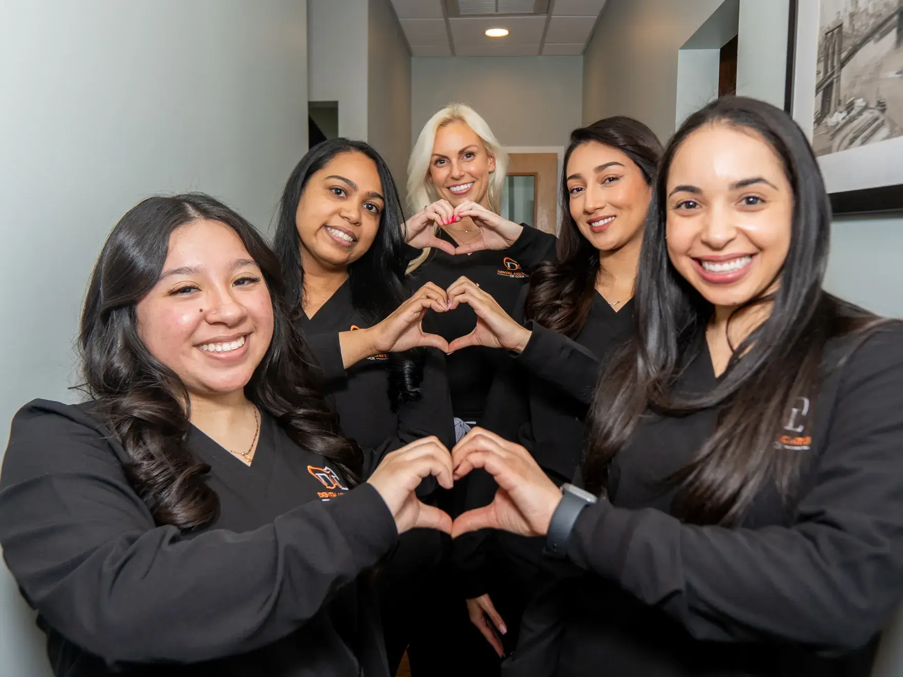Five women in black uniforms making heart shapes with their hands, smiling in a hallway.