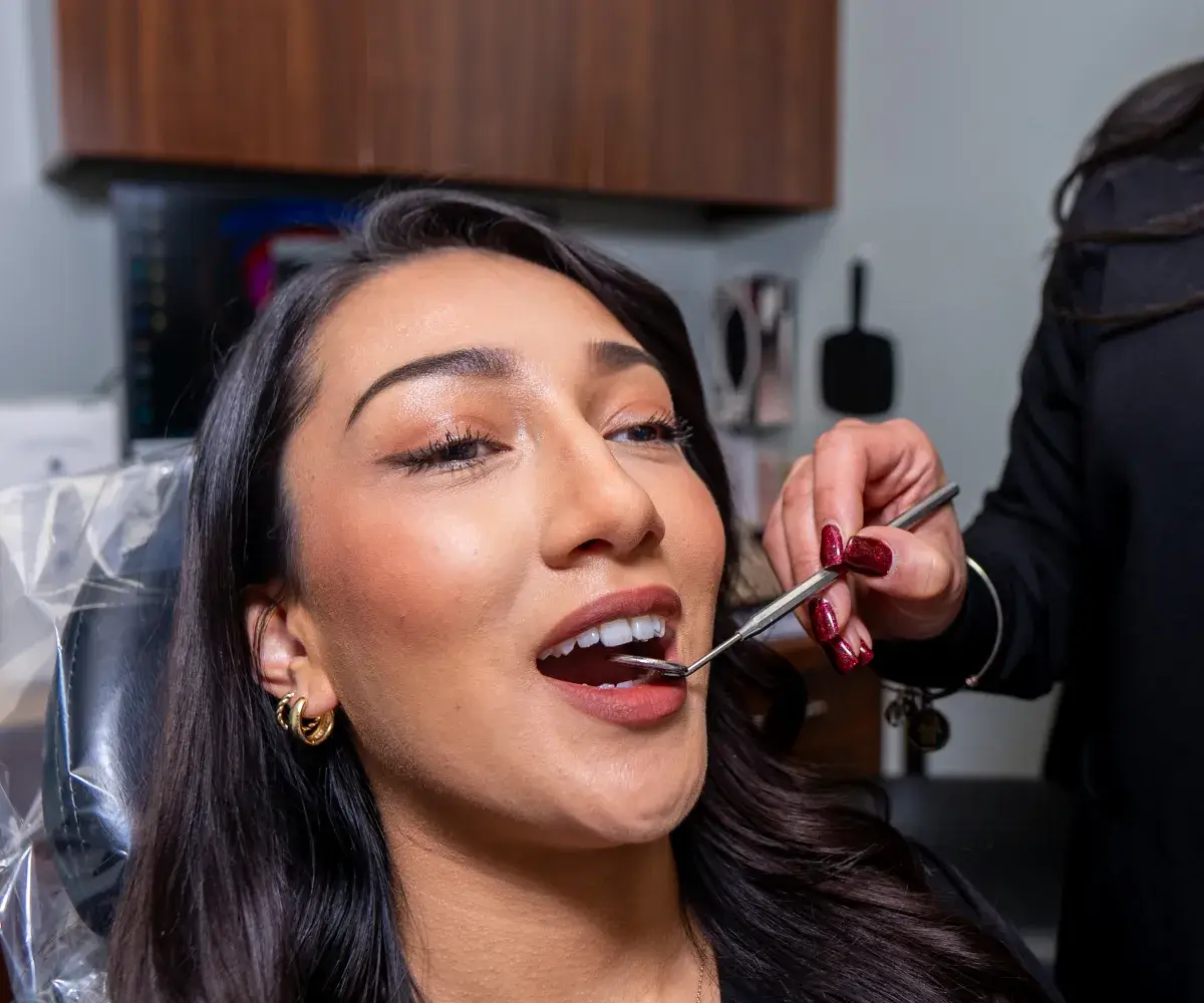 Woman with dark hair reclining in dental chair while dentist examines her teeth using a dental mirror.