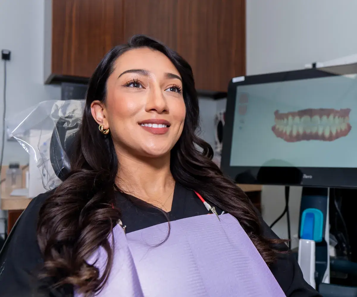 A woman sits in a dental chair, smiling, with a computer displaying dental images behind her.