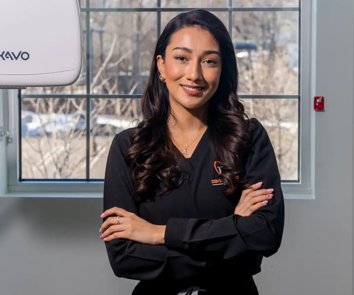 A woman in a black shirt stands with arms crossed, smiling in front of a window.