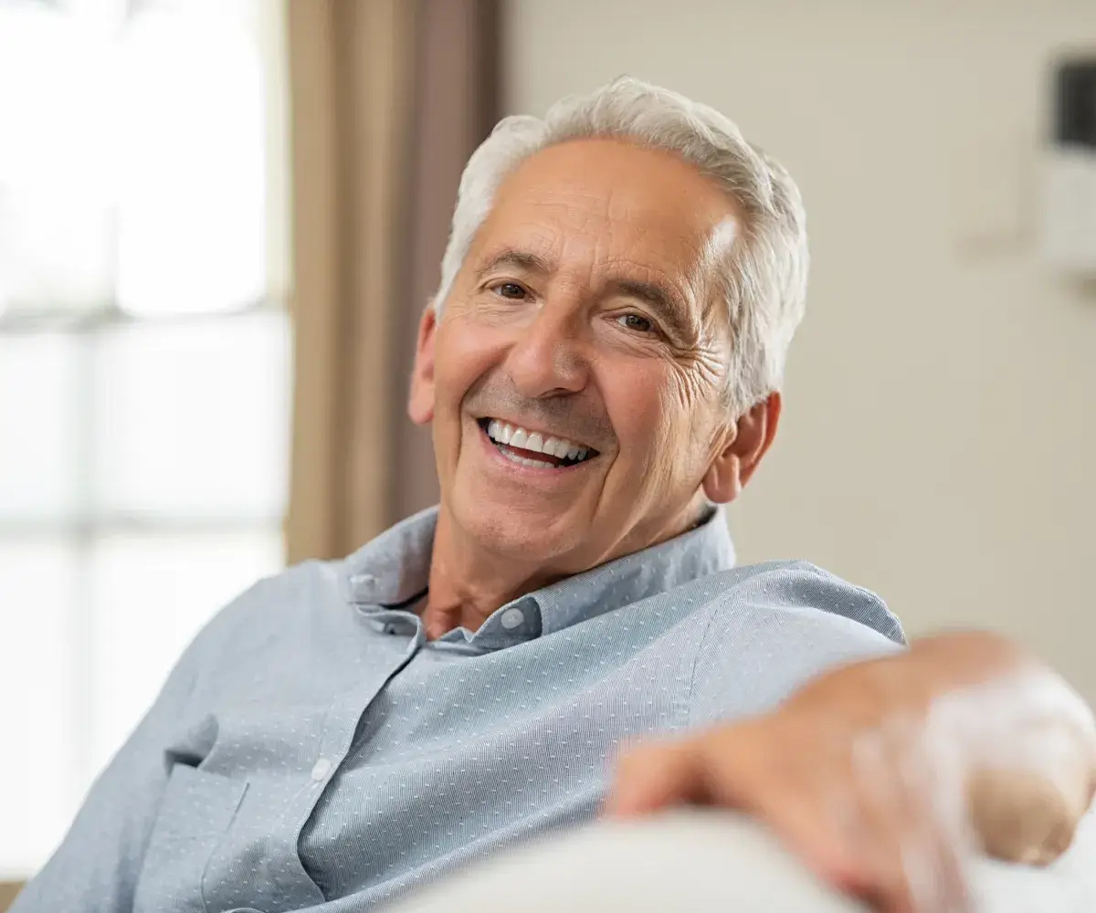 Smiling older man in a light blue shirt sitting indoors, looking at the camera.
