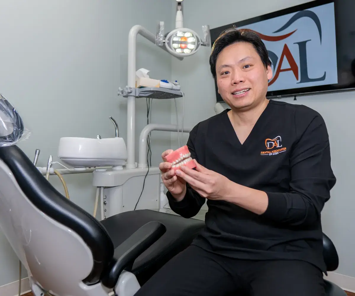 Smiling male dentist in black scrubs holding a dental model in a dental clinic.
