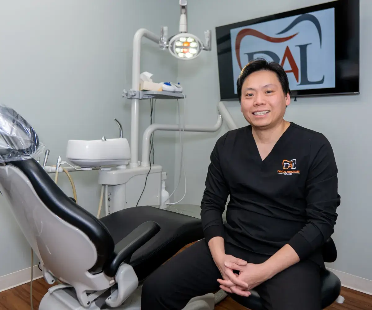 A dentist in scrubs sits on a chair in a dental office, smiling at the camera.