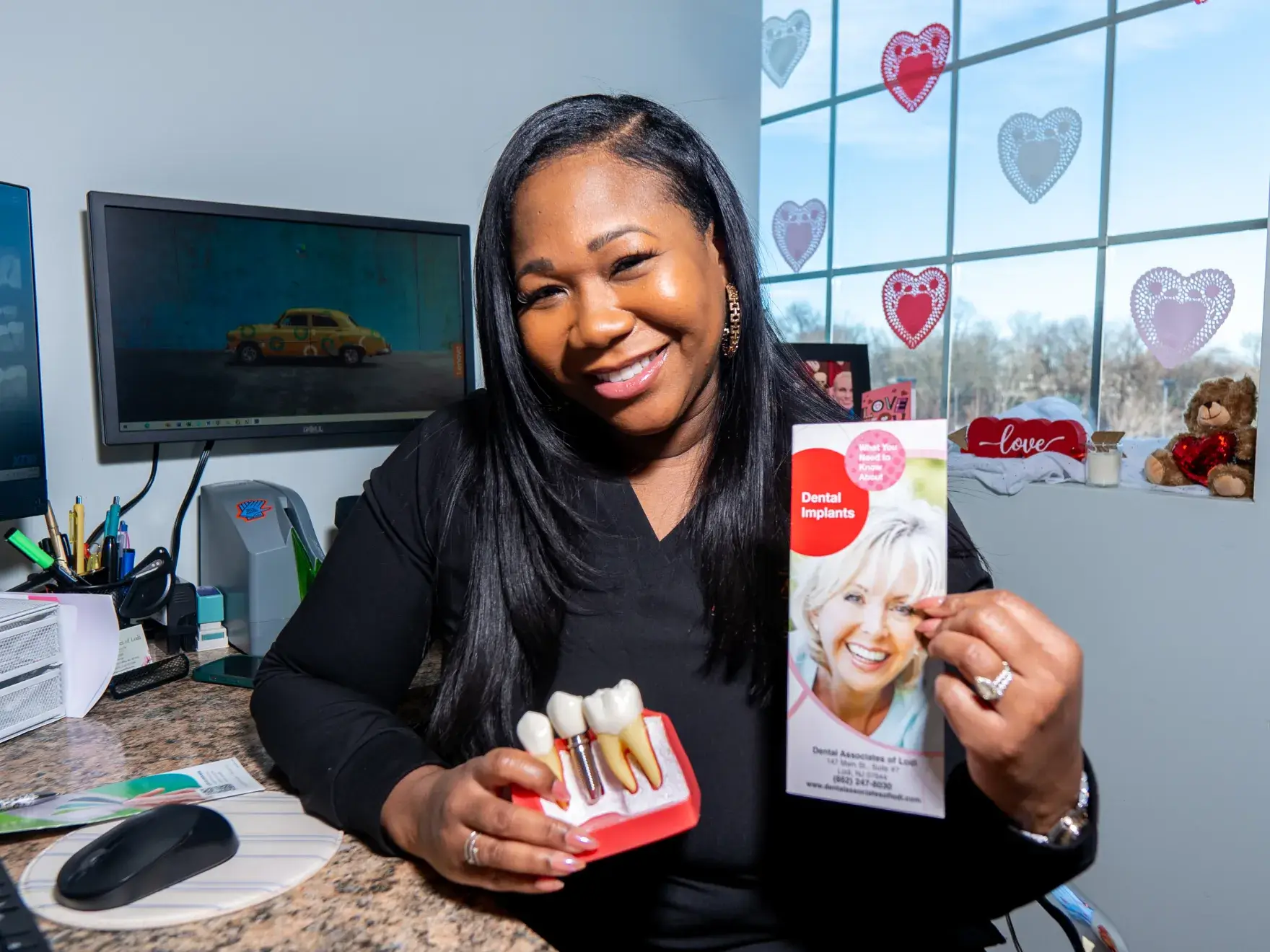 A smiling woman holds a dental implant model and a brochure titled "Dental Implants" in an office.