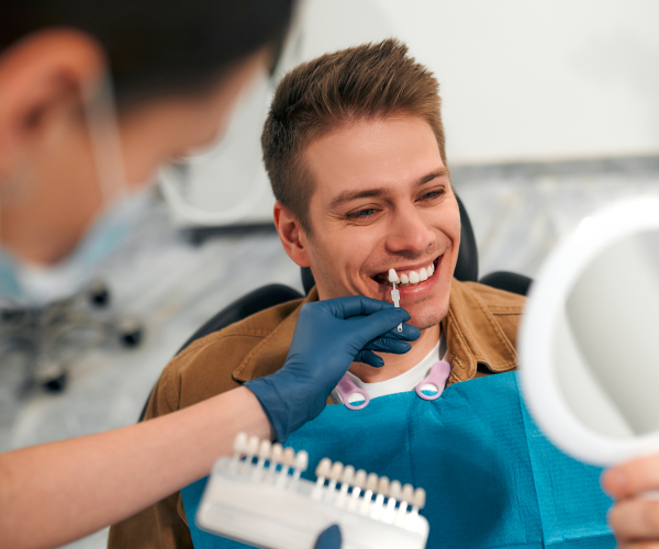 A dentist holds a tooth color chart near a smiling patient holding a small mirror.