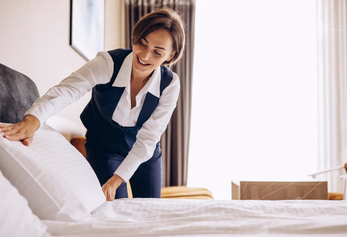 Smiling woman in uniform making a bed in a sunlit hotel room.