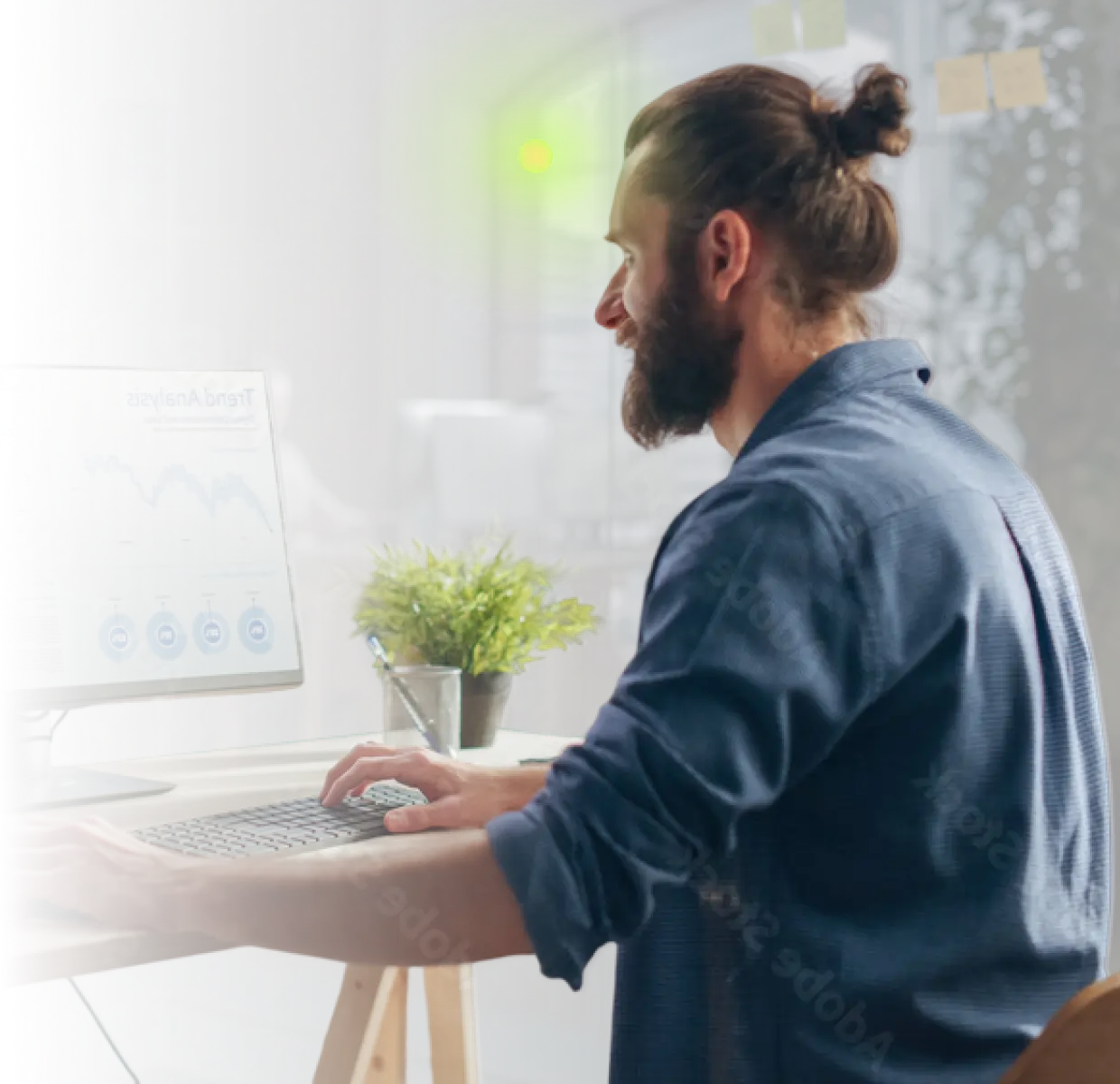 Man with beard and bun works on a computer with graphs visible on the screen in an office.